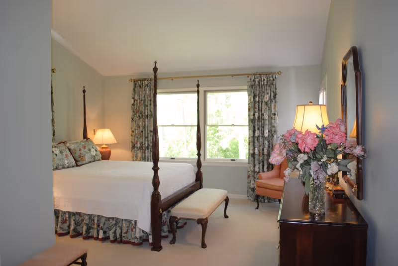A bright bedroom with a large four-poster bed covered in white bedding and floral pillows. There is a matching floral bed skirt and curtains on the window behind the bed. A wooden bench with a cushioned seat is placed at the foot of the bed. To the right, there is a wooden dresser with a large mirror above it, a table lamp, and a vase of pink flowers. An upholstered armchair is positioned near the window.