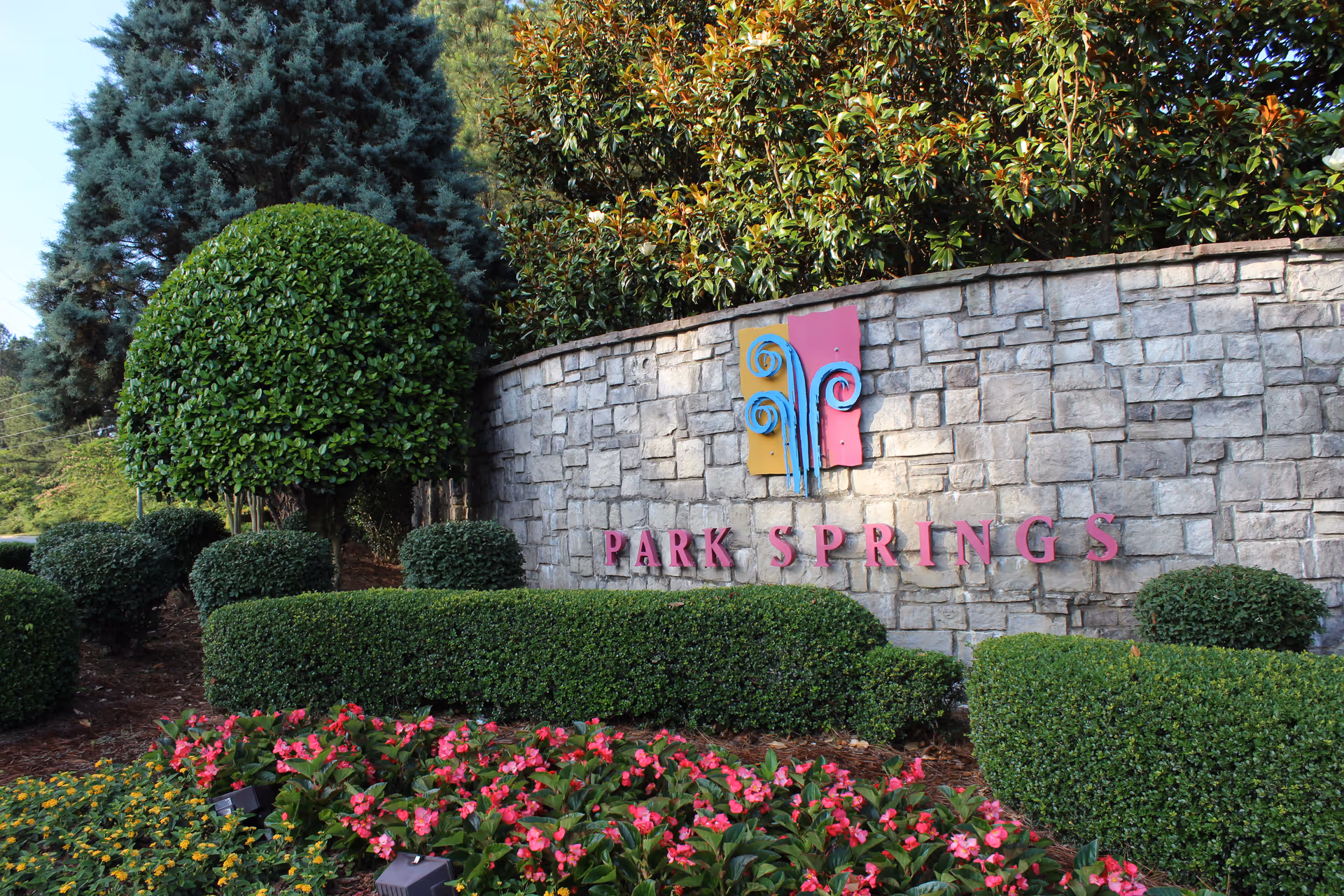 Stone wall with the colorful Park Springs logo and name mounted on it, surrounded by neatly trimmed bushes and vibrant flower beds, with tall trees in the background under a clear sky.
