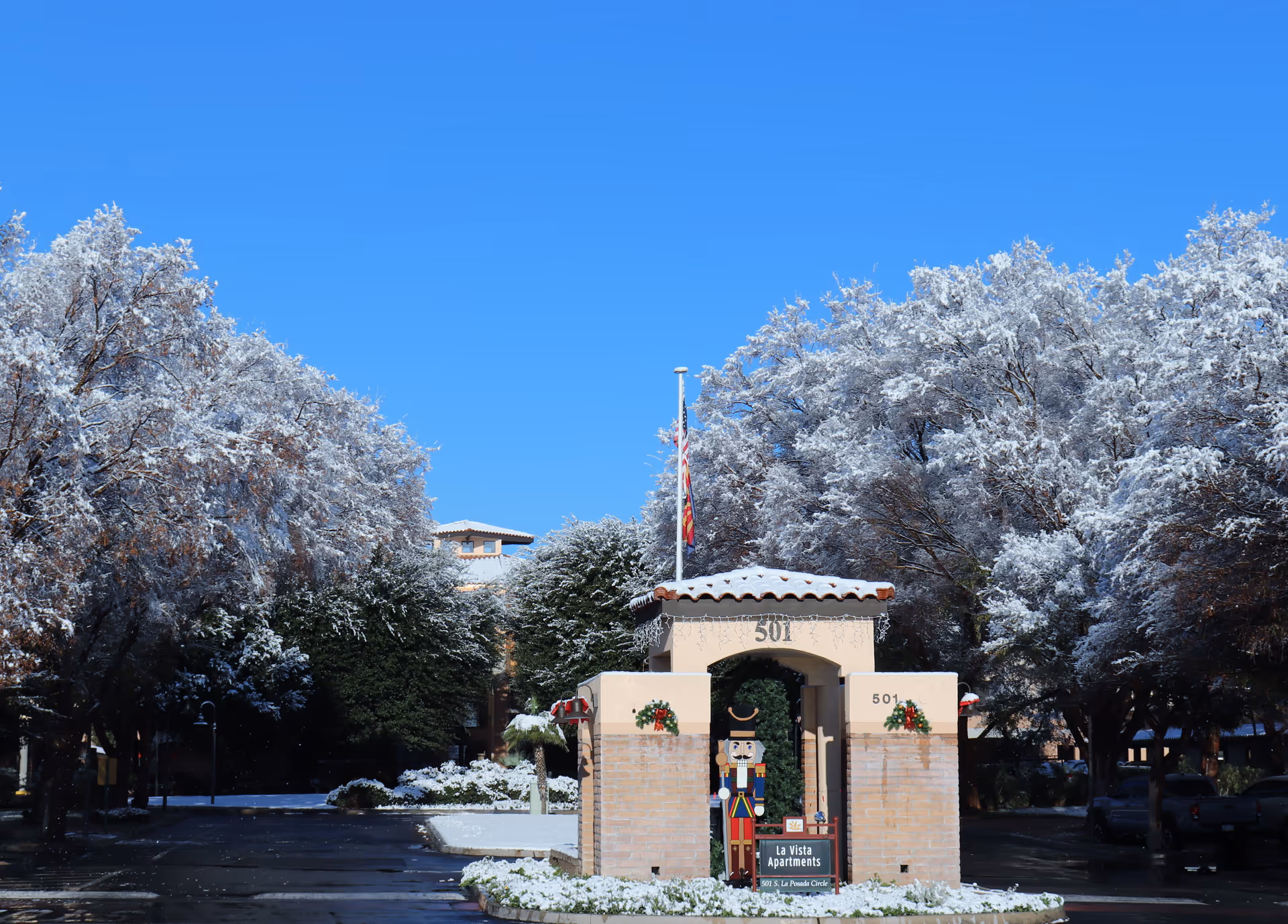 Snow-covered trees frame the entrance archway of a residential building marked '501' with holiday wreaths, a nutcracker figure and an American flag under a clear blue sky.