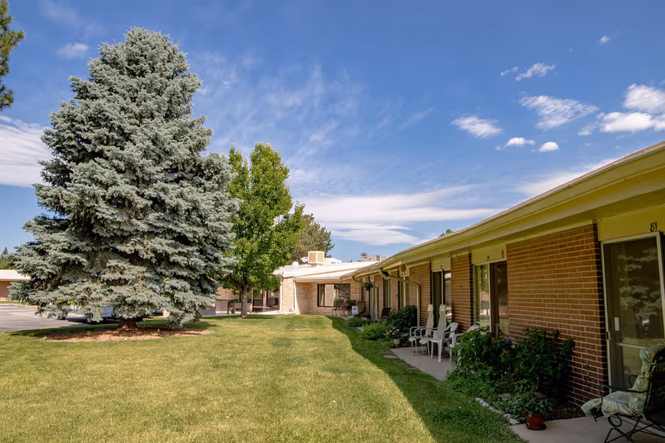 Outdoor view of a senior living facility with a large green lawn, several trees including a tall pine tree, and a single-story brick building with patios and chairs outside the rooms under a blue sky with some clouds.