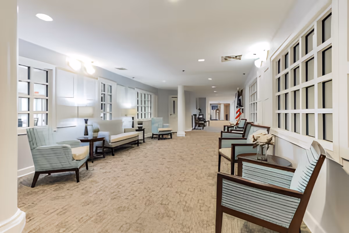 A long, well-lit hallway with multiple seating areas featuring armchairs and small tables along both sides. The walls have large windows with white frames, and there are lamps providing additional lighting. The carpet is beige, and the overall atmosphere is calm and inviting.