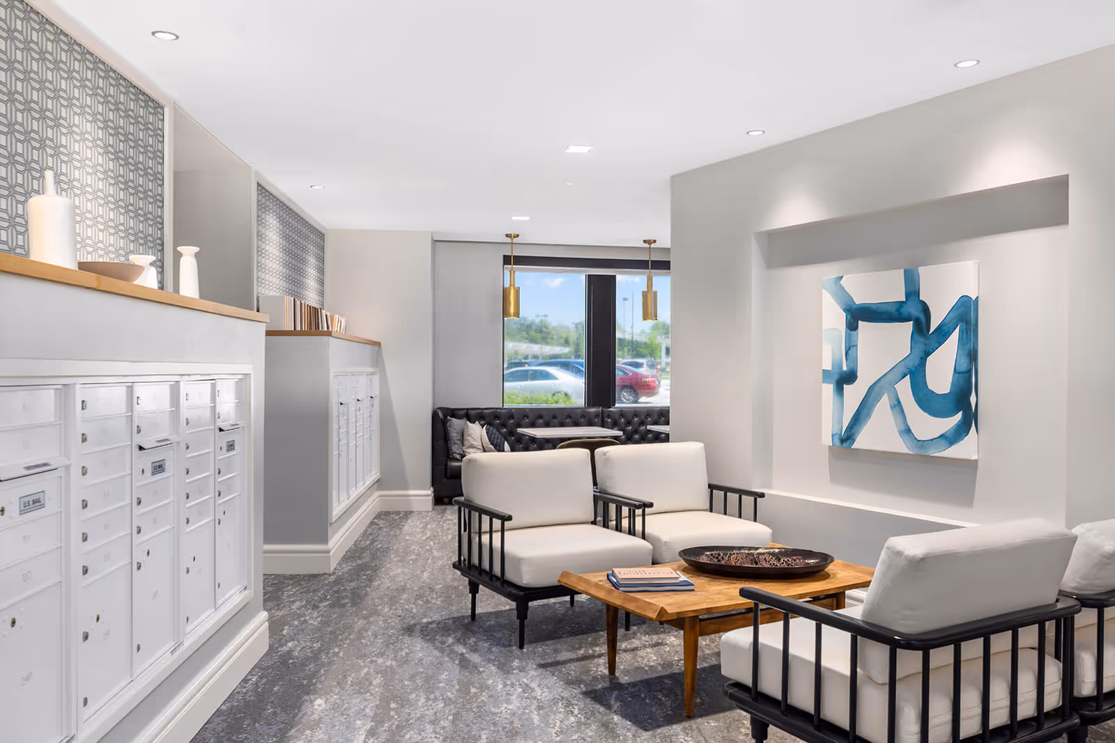 A modern interior common area featuring a seating arrangement with white cushioned chairs and a wooden coffee table. Behind the seating area, there is a black tufted bench with pillows near a window showing parked cars outside. On the left side, there are white mailboxes built into the wall with decorative vases on top. The walls are light gray with a blue abstract painting on the right wall and patterned wallpaper above the mailboxes.