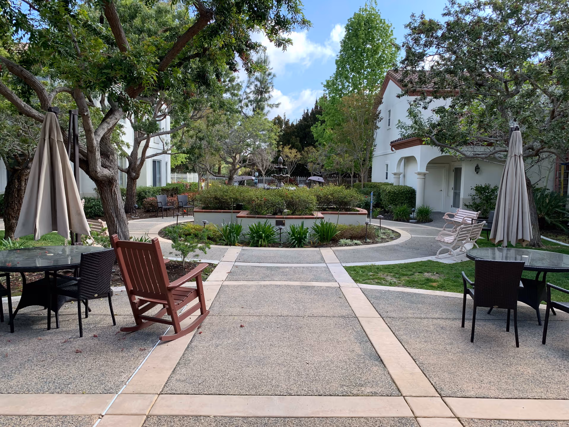 Outdoor patio area with round glass tables, chairs, and umbrellas. There is a red wooden rocking chair in the foreground. The area is surrounded by trees, bushes, and a circular garden bed with a fountain in the center. White buildings with red-tiled roofs are visible in the background under a partly cloudy sky.
