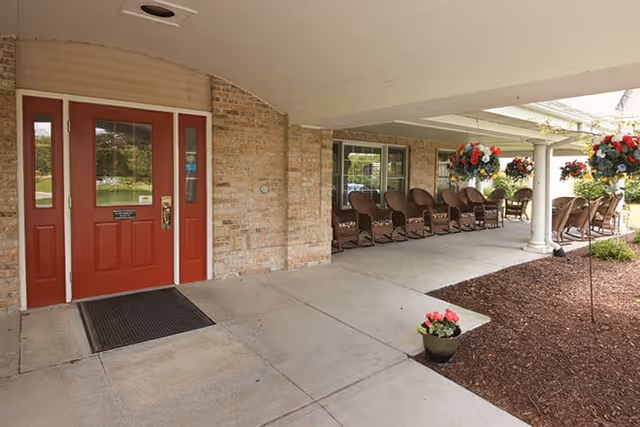 Covered outdoor entrance area of a senior living facility with a red double door, brick walls, and a row of brown rocking chairs along the wall. There are hanging flower baskets and a small potted plant near the walkway.