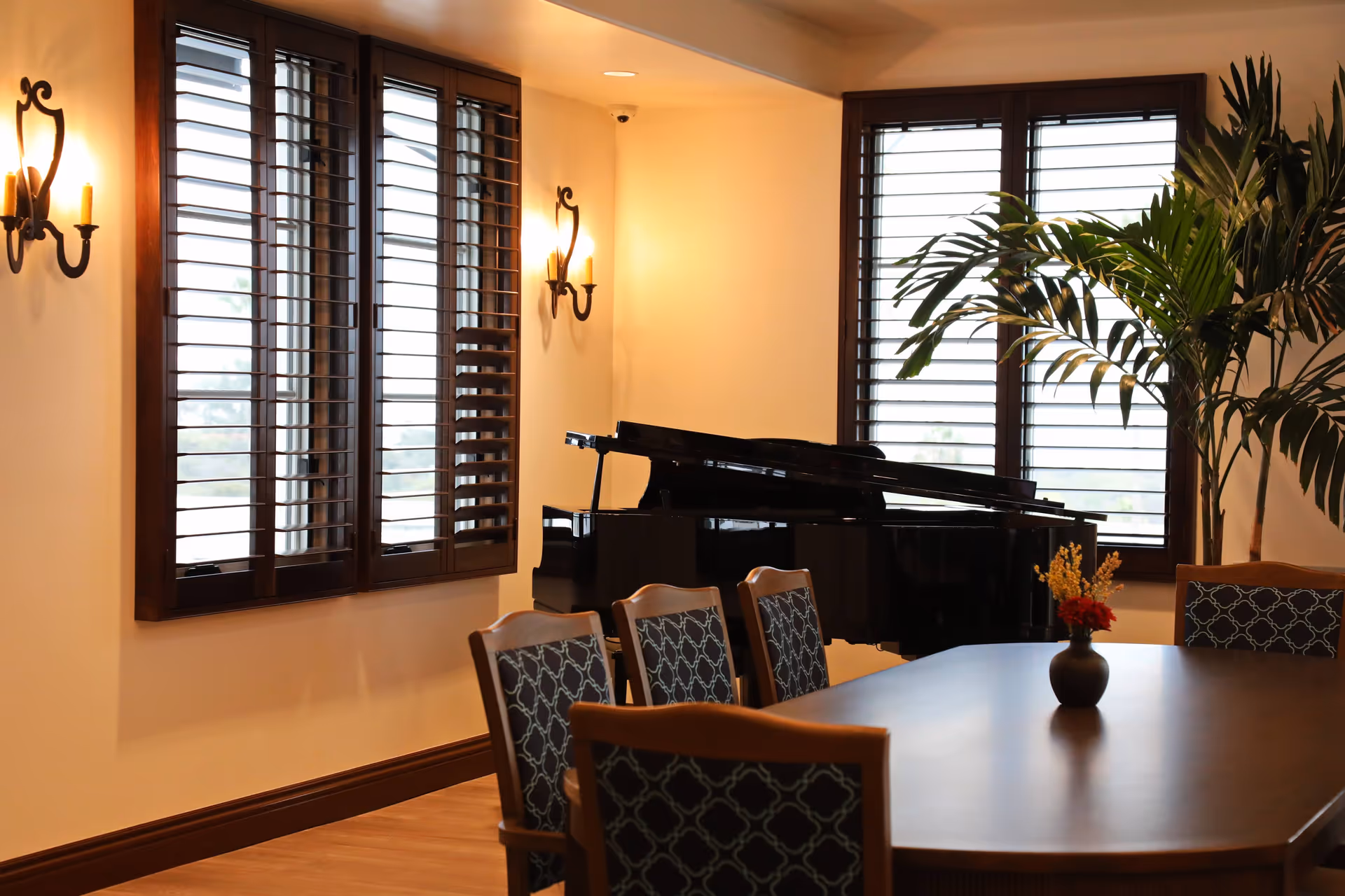 A cozy interior room with a wooden table surrounded by chairs with patterned upholstery. A black grand piano is positioned near windows with wooden shutters. Two wall-mounted candle-style light fixtures illuminate the room, and a large potted plant is placed near the piano.