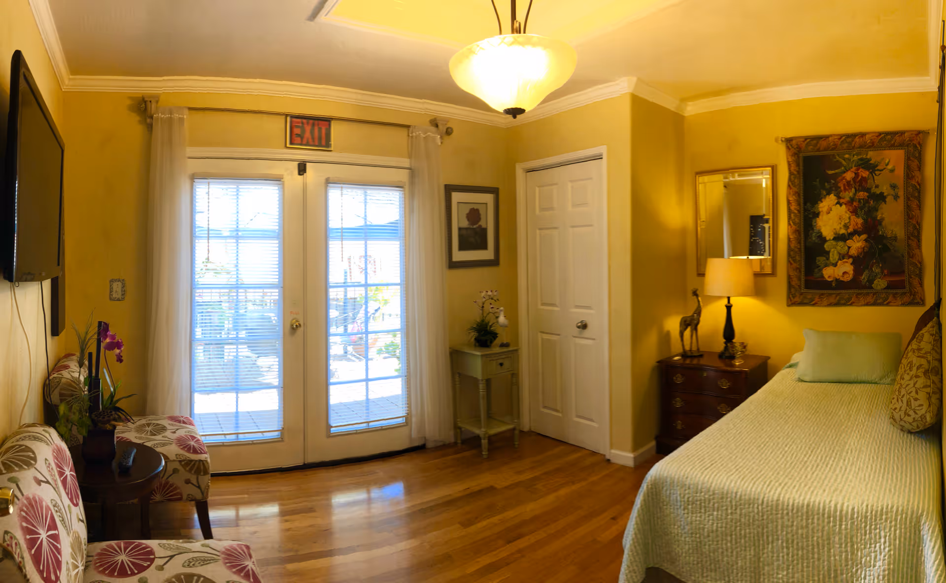 A cozy room with yellow walls featuring a single bed with a light green bedspread and pillows, a wooden nightstand with a lamp and decorative giraffe figurine, a framed floral painting, a mirror, and a small table with a plant. There are two patterned chairs and a small round table near glass double doors with blinds leading outside. The floor is wooden, and a ceiling light fixture is visible.