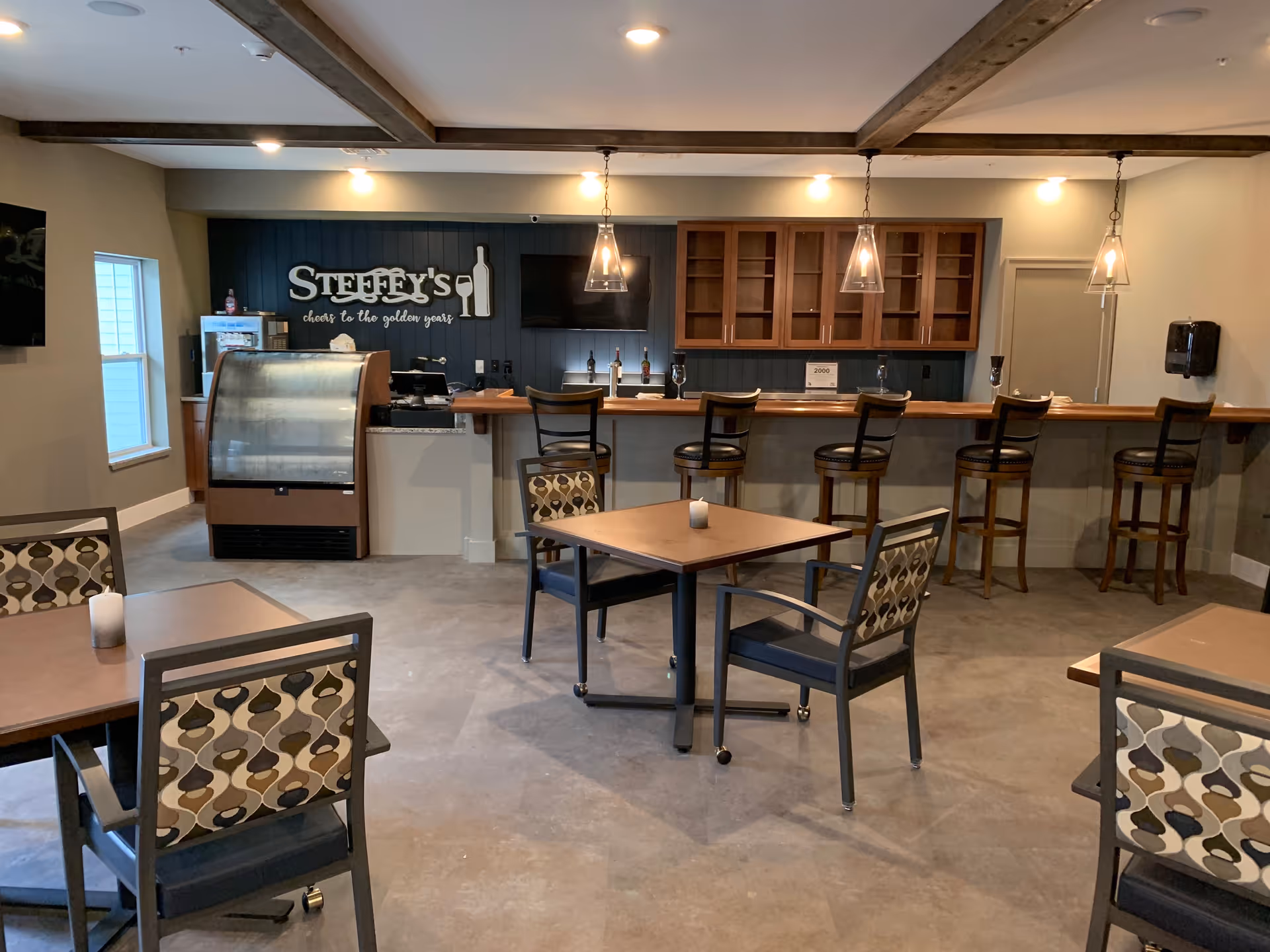 Interior view of a small bar and seating area in a retirement living facility. The bar has five high stools and a wooden countertop with three pendant lights hanging above. Behind the bar are wooden cabinets and a sign that reads 'STEFFEY'S cheers to the golden years'. There are several tables with patterned chairs in the foreground, and a small refrigerated display case near the bar.