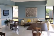 A bright room with light blue walls featuring a beige couch, a coffee table with yellow flowers, and several wooden chairs around tables. There is a flat-screen TV mounted on the wall to the left and large windows letting in natural light.