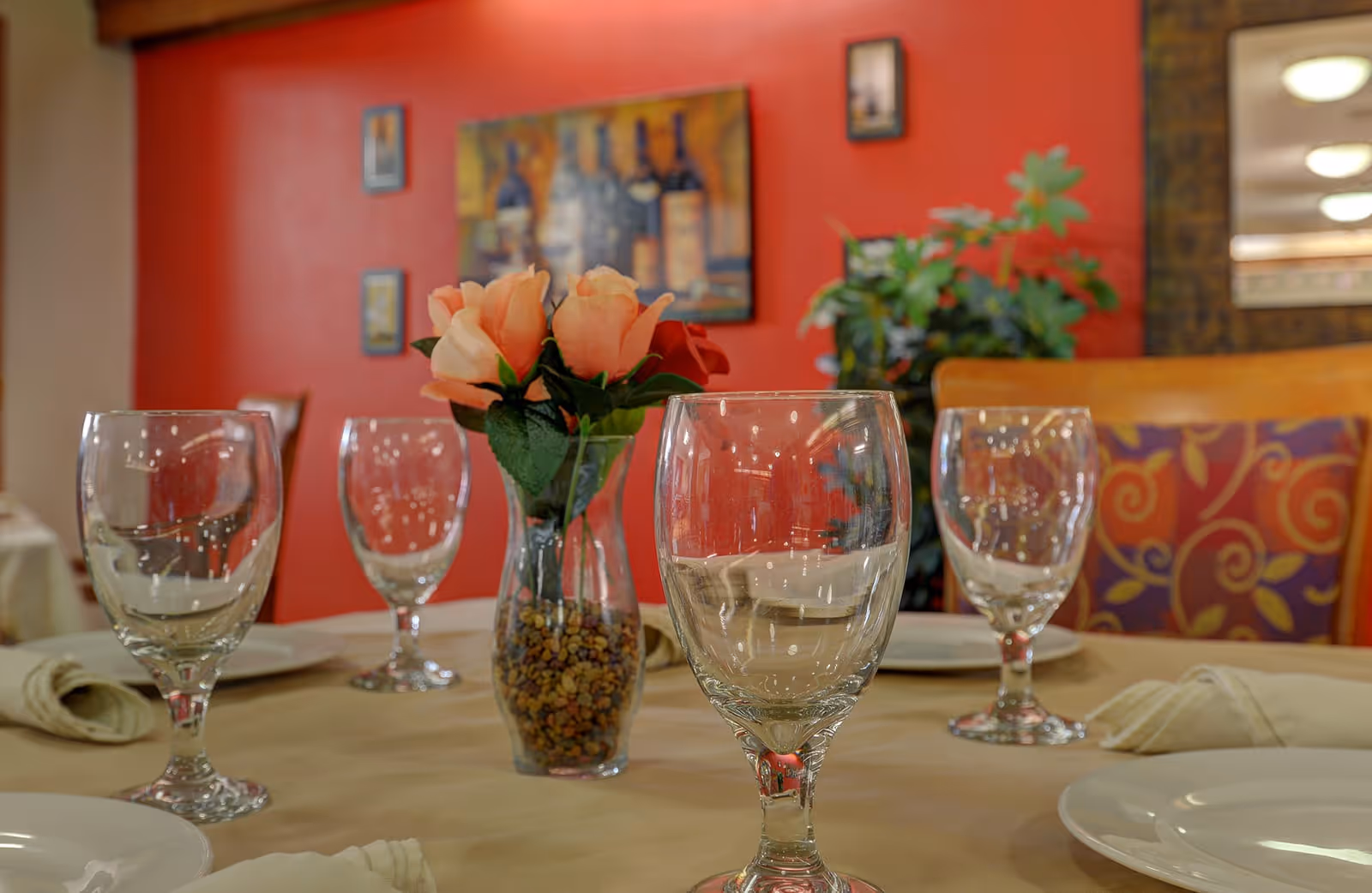 Close-up view of a dining table set with empty water glasses, white plates, and beige napkins. A small vase with peach-colored artificial flowers and decorative stones is placed in the center. The background features a red wall with framed artwork and a plant next to a wooden chair with a colorful cushion.