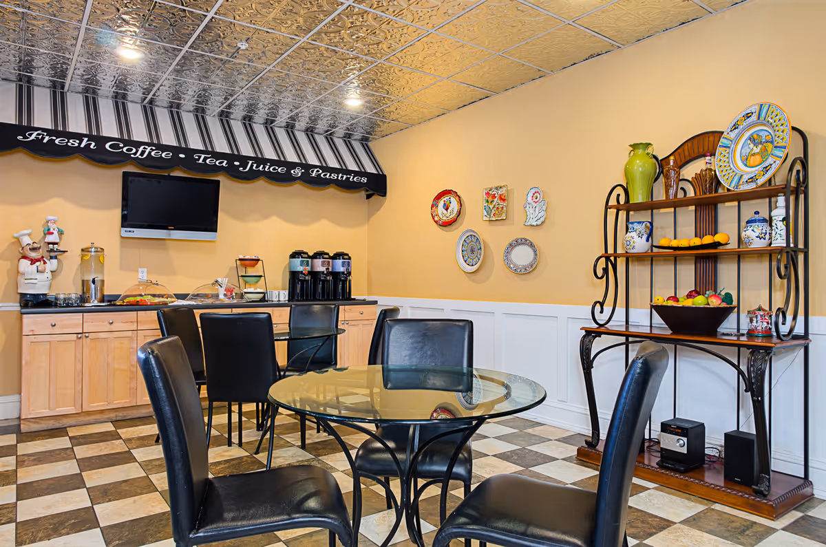 A cozy dining area with a round glass table surrounded by black leather chairs. Against the wall, there is a counter with coffee dispensers, a water dispenser, and a display of pastries. Above the counter is a black and white striped awning with the words 'Fresh Coffee • Tea • Juice & Pastries'. A TV is mounted on the wall above the counter. On the right side, a decorative shelving unit holds colorful plates, vases, and bowls of fruit. The floor has a checkered tile pattern and the ceiling features a decorative metallic design.