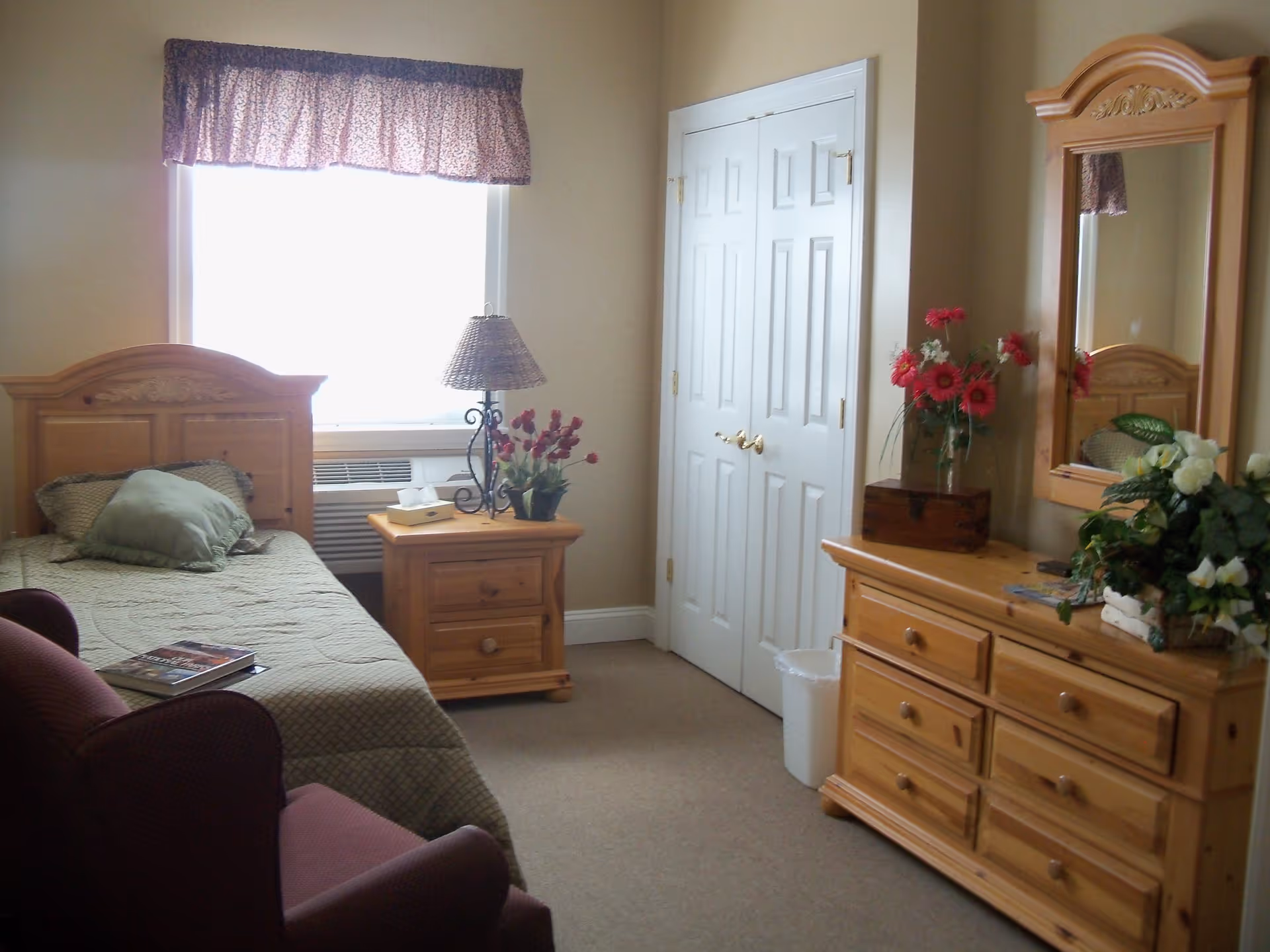 A cozy bedroom in an assisted living facility featuring a single wooden bed with green bedding and pillows, a wooden nightstand with a lamp and a flower pot, a wooden dresser with a mirror adorned with flower arrangements, a white double-door closet, and a maroon armchair.