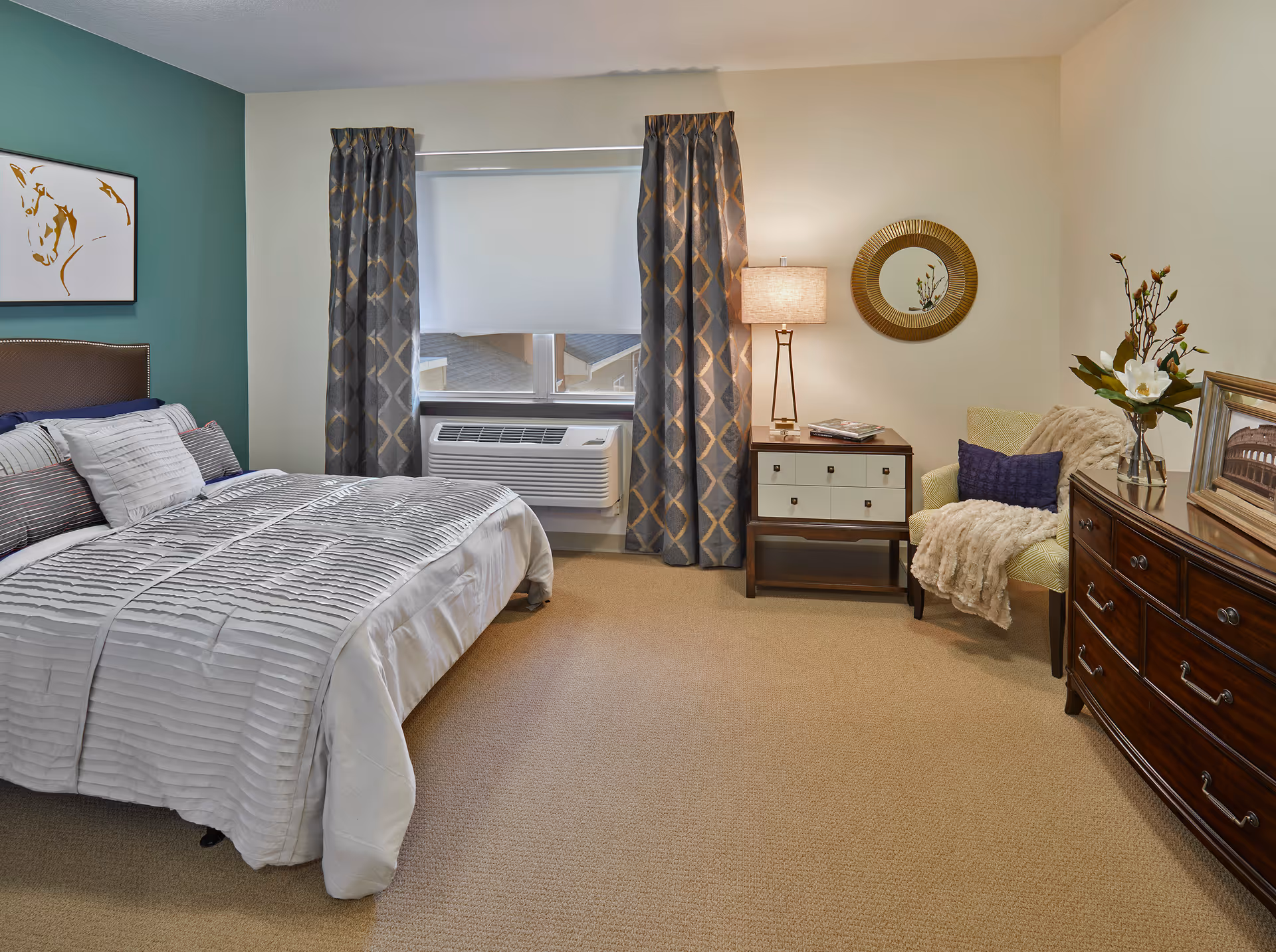 A cozy bedroom featuring a neatly made bed with white bedding and multiple pillows, a window with patterned curtains and a white roller shade, an air conditioning unit below the window, a wooden nightstand with a lamp and books, a comfortable armchair with a throw blanket and pillow, a round decorative mirror on the wall, and a wooden dresser with a framed picture and a vase with flowers.