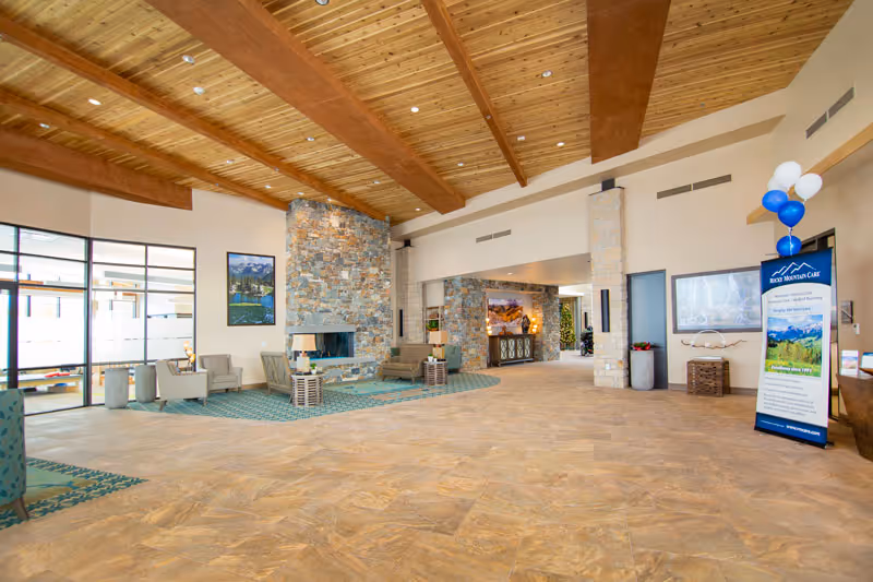 Spacious and well-lit common area with a high wooden beam ceiling, stone fireplace, seating arrangements with chairs and tables, and large windows allowing natural light. There is a standing banner with balloons on the right side near the entrance.