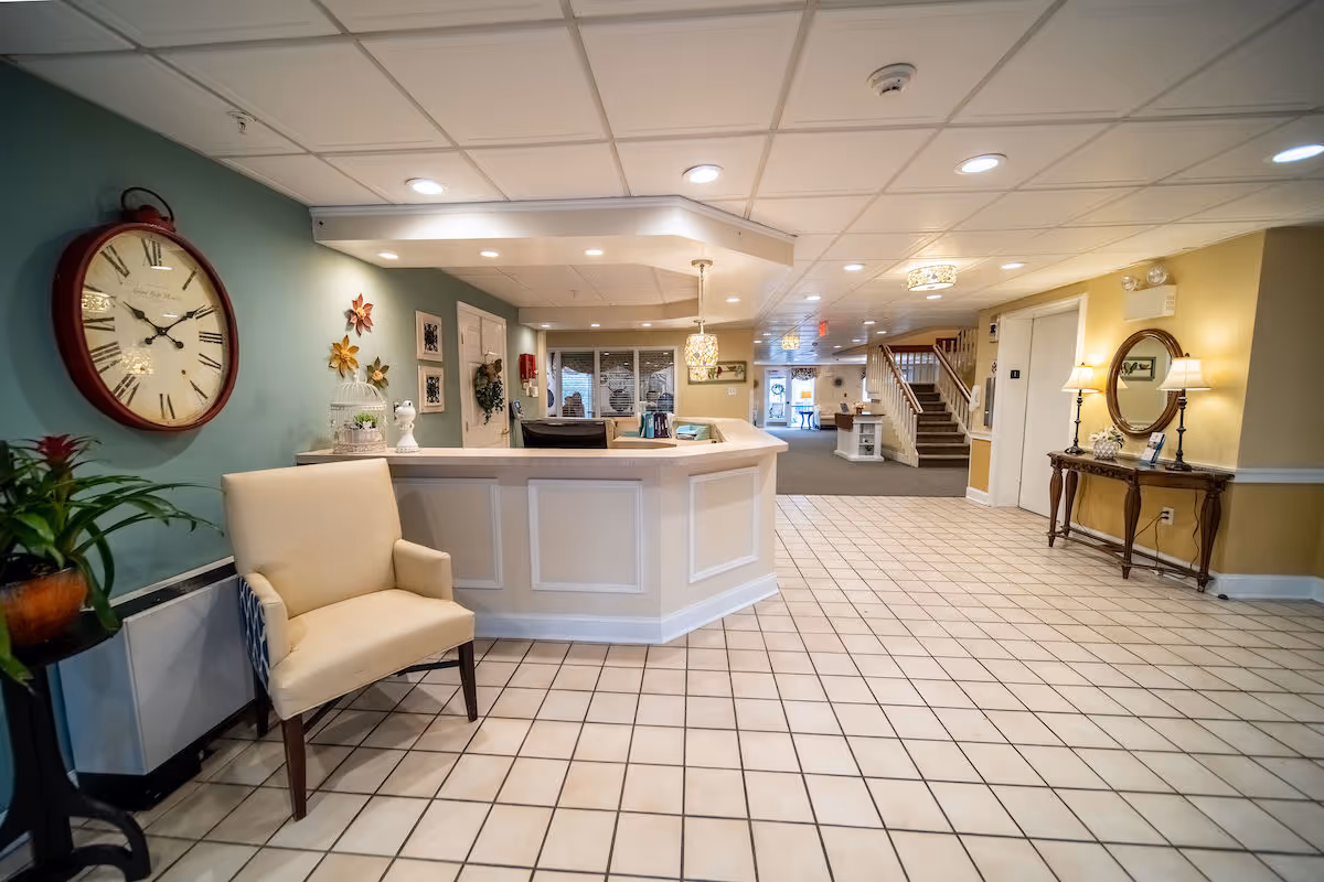Reception area of a senior living facility with a white reception desk, beige tiled floor, a beige armchair, a large wall clock, decorative plants, and a hallway leading to stairs and other rooms.
