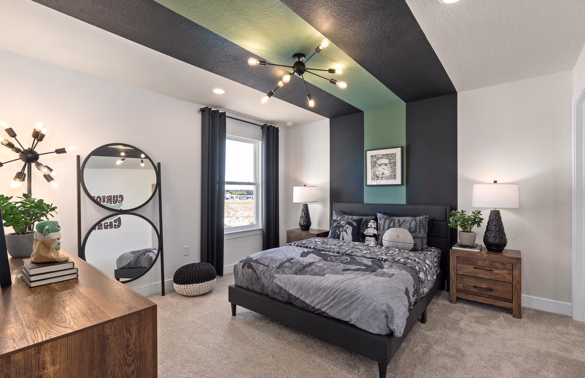 A modern bedroom with a black and green accent wall behind a bed with gray Star Wars-themed bedding. The room features two wooden nightstands with black lamps, a wooden dresser with decorative items, a round double mirror, a window with black curtains, and a beige carpeted floor.