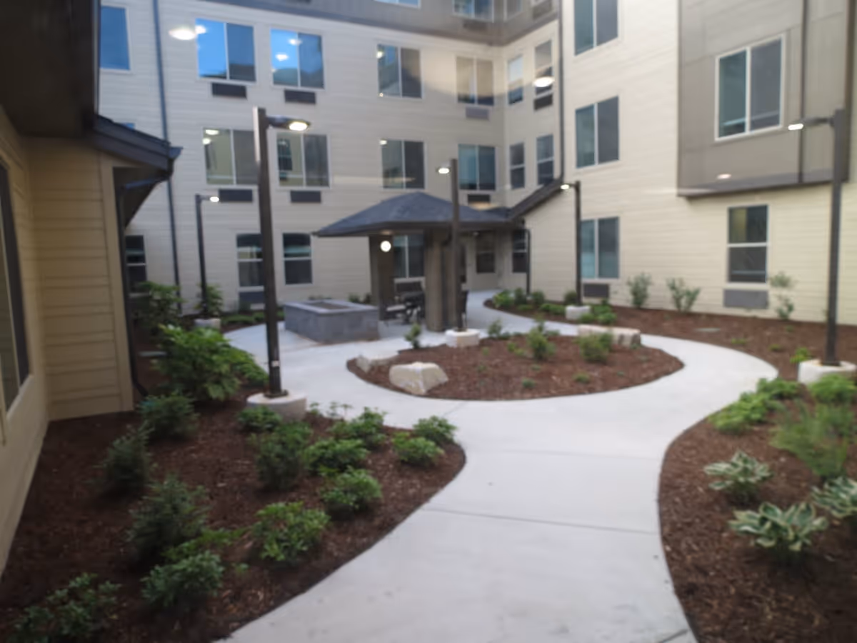 Outdoor courtyard area of a senior living facility with a winding concrete pathway, landscaped garden beds with small shrubs, a covered seating area with a roof, and multiple windows of the surrounding building visible.
