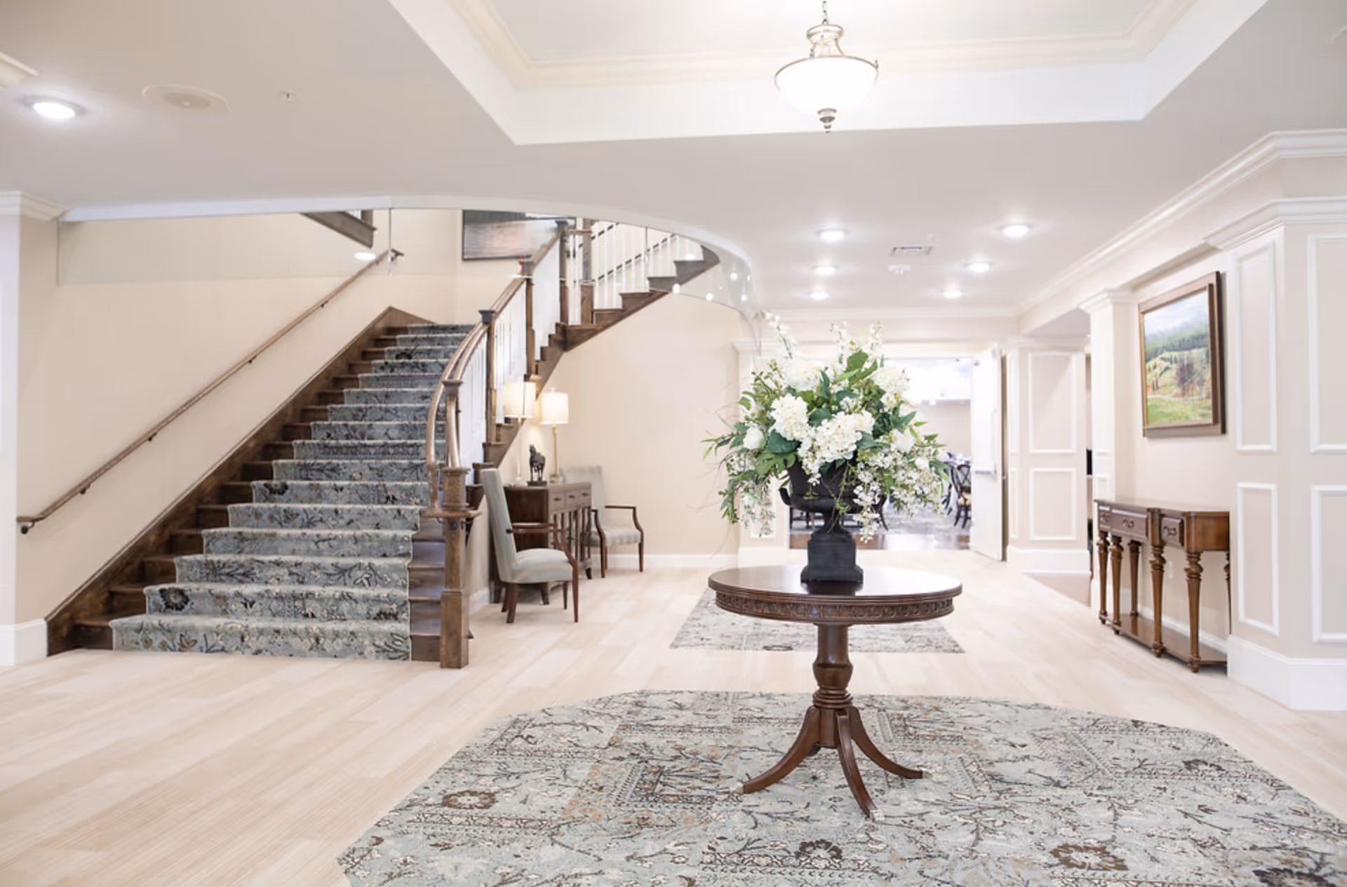 Bright and elegant interior space featuring a curved staircase with a patterned carpet runner, a round wooden table with a large floral arrangement in the center, light wood flooring, and classic furniture including chairs and side tables. The walls are adorned with molding and a framed landscape painting, and the area is well-lit with ceiling lights and a hanging light fixture.