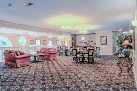 Spacious carpeted senior living common area with upholstered sofas, round dining tables and chairs, chandeliers, and a balcony railing overlooking large arched windows.