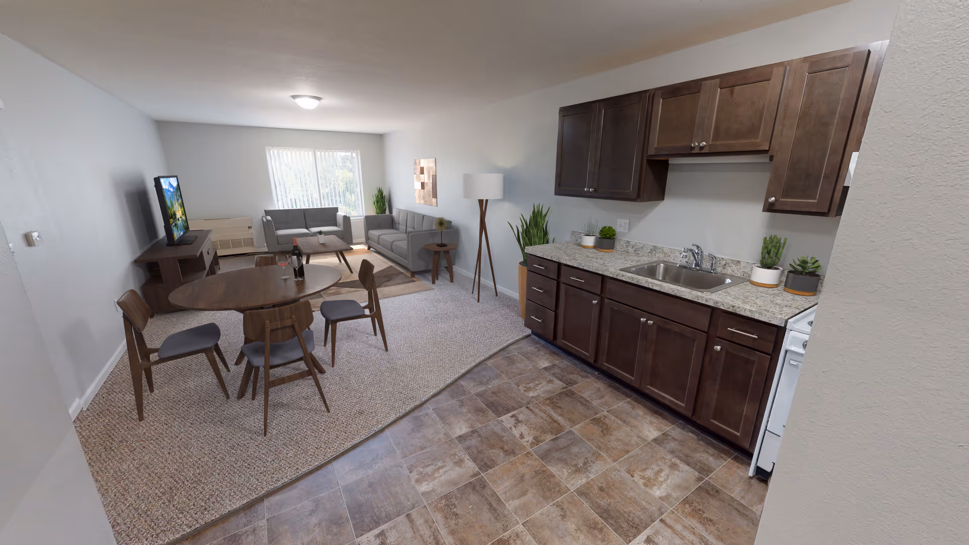 Open-plan apartment interior with a kitchenette featuring dark cabinets and a sink, a round dining table, and a living area with gray sofas and a TV.