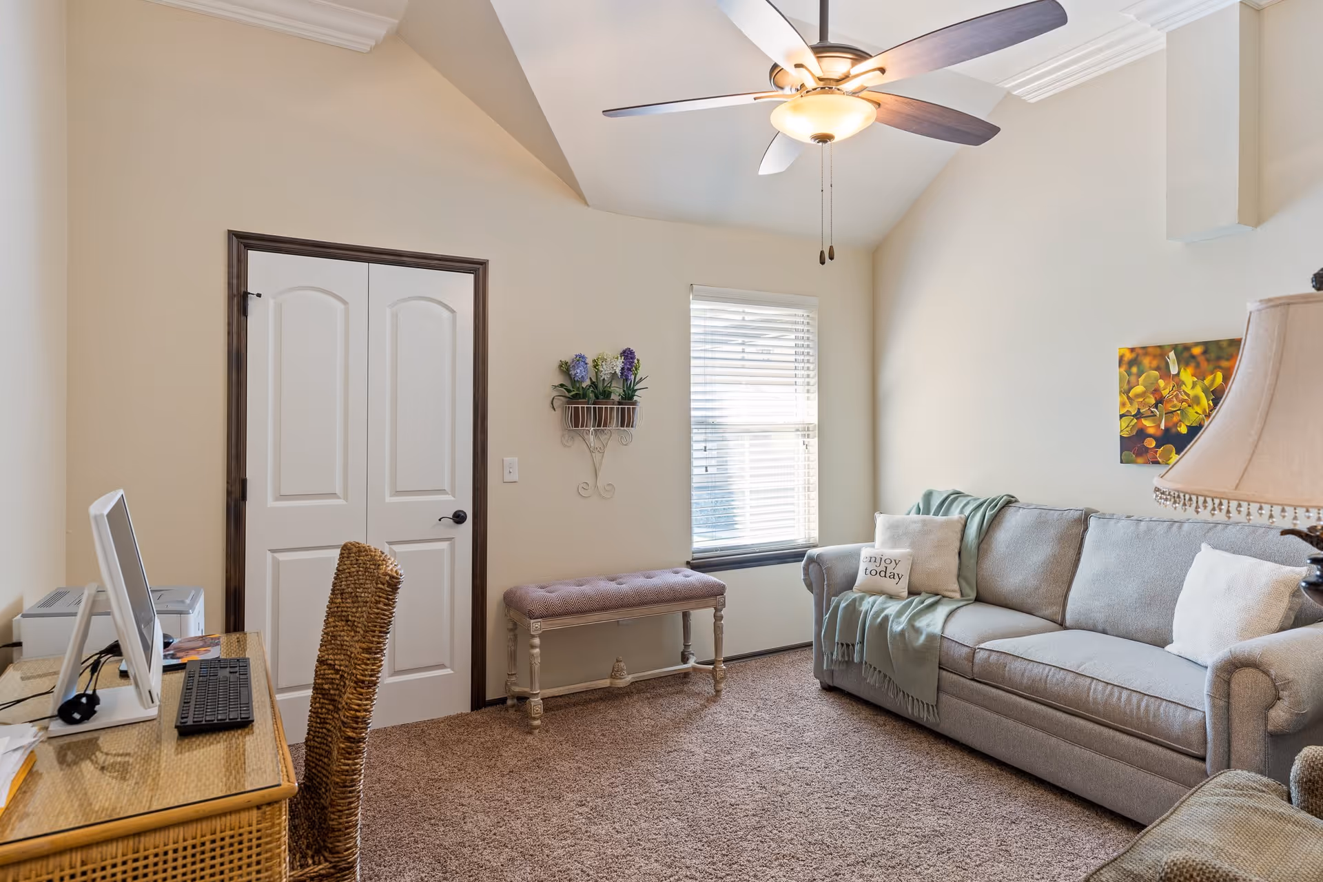 A cozy living room with a beige carpet, a gray sofa adorned with pillows and a green throw blanket, a wicker desk with a computer and keyboard, a wicker chair, a small cushioned bench under a window with blinds, a ceiling fan with lights, and a wall decoration with flowers.