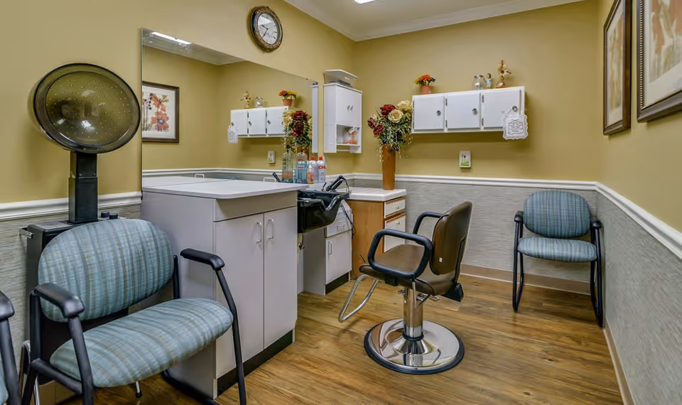 Interior of a senior living facility hair salon with a hair washing station, a salon chair, two waiting chairs, a large mirror, wall cabinets, and decorative flowers in a vase. The walls are painted yellow with white wainscoting and the floor is wood.
