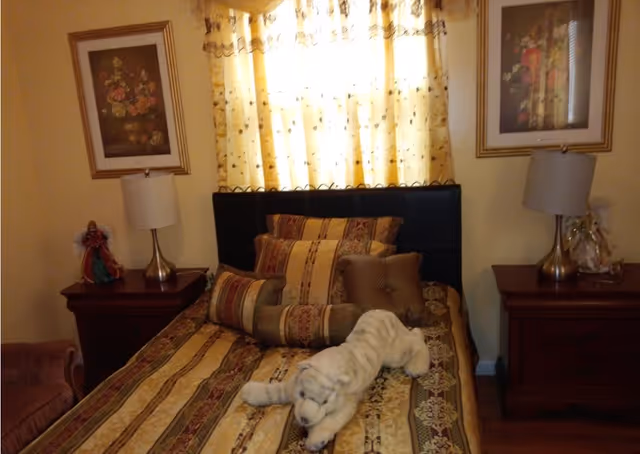 A bedroom with a bed covered in a patterned bedspread and multiple decorative pillows. A white stuffed animal resembling a tiger is placed on the bed. There are two wooden nightstands on either side of the bed, each with a lamp and decorative items. The window behind the bed has yellow curtains with a floral pattern, and framed floral artwork hangs on the walls above each nightstand.