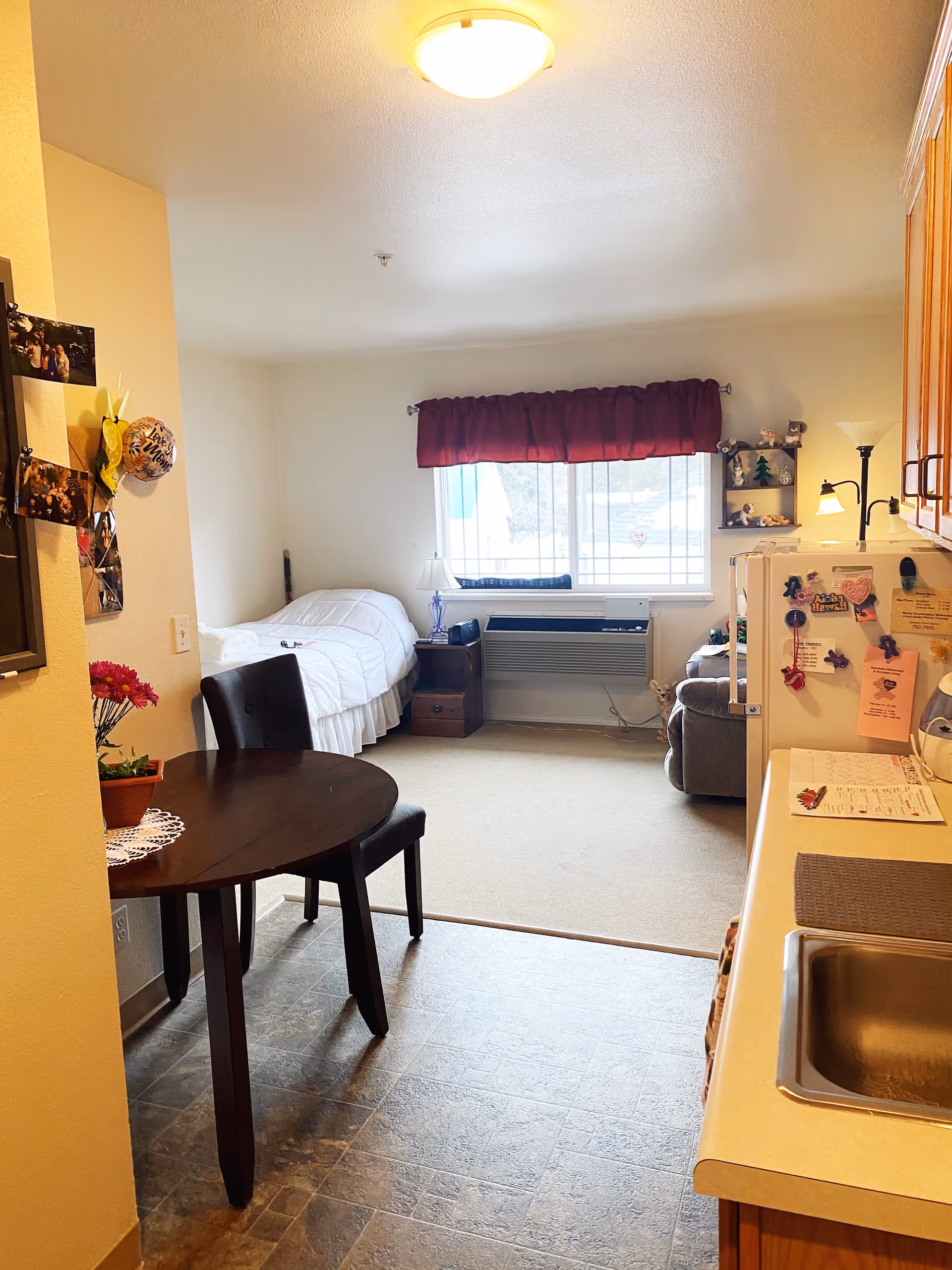 Interior view of a small retirement living space featuring a kitchenette with a sink and refrigerator on the right, a round dining table with two chairs on the left, and a bed with white bedding near a window with a red valance. A small nightstand with a lamp is next to the bed, and a cozy armchair is partially visible near the window. The floor transitions from tiled in the kitchen area to carpeted in the living and sleeping area.
