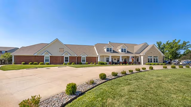 Single-story brick and siding senior living facility with a covered entrance, curved driveway, and landscaped lawn under a clear blue sky.