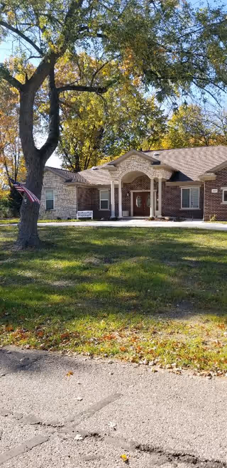 Front exterior view of a single-story brick and stone building with a covered entrance supported by columns. There is a large tree with an American flag attached to it in the grassy yard in front of the building. The sky is clear and the surrounding trees have autumn foliage.