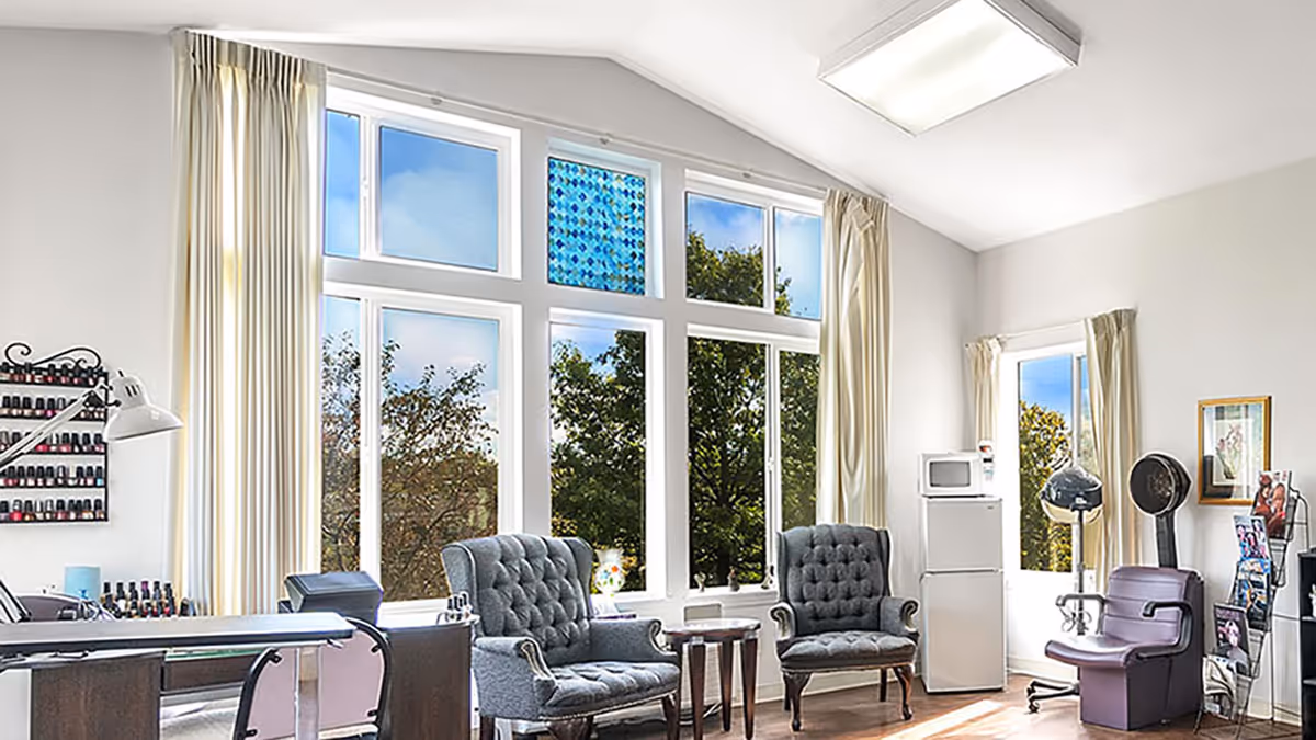 Bright interior common room with tall windows, two tufted armchairs, salon equipment and a nail polish display.