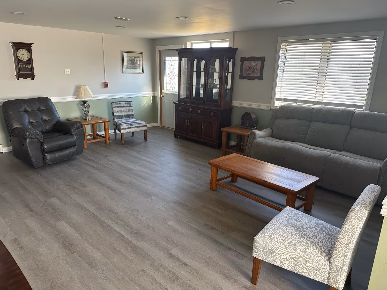 A living room with light gray walls and wood flooring. The room contains a gray sofa, a black leather recliner, a patterned armchair, a wooden coffee table, two wooden side tables with lamps, a wooden china cabinet, a wall clock, and framed pictures. There is a window with blinds and a door with decorative glass.