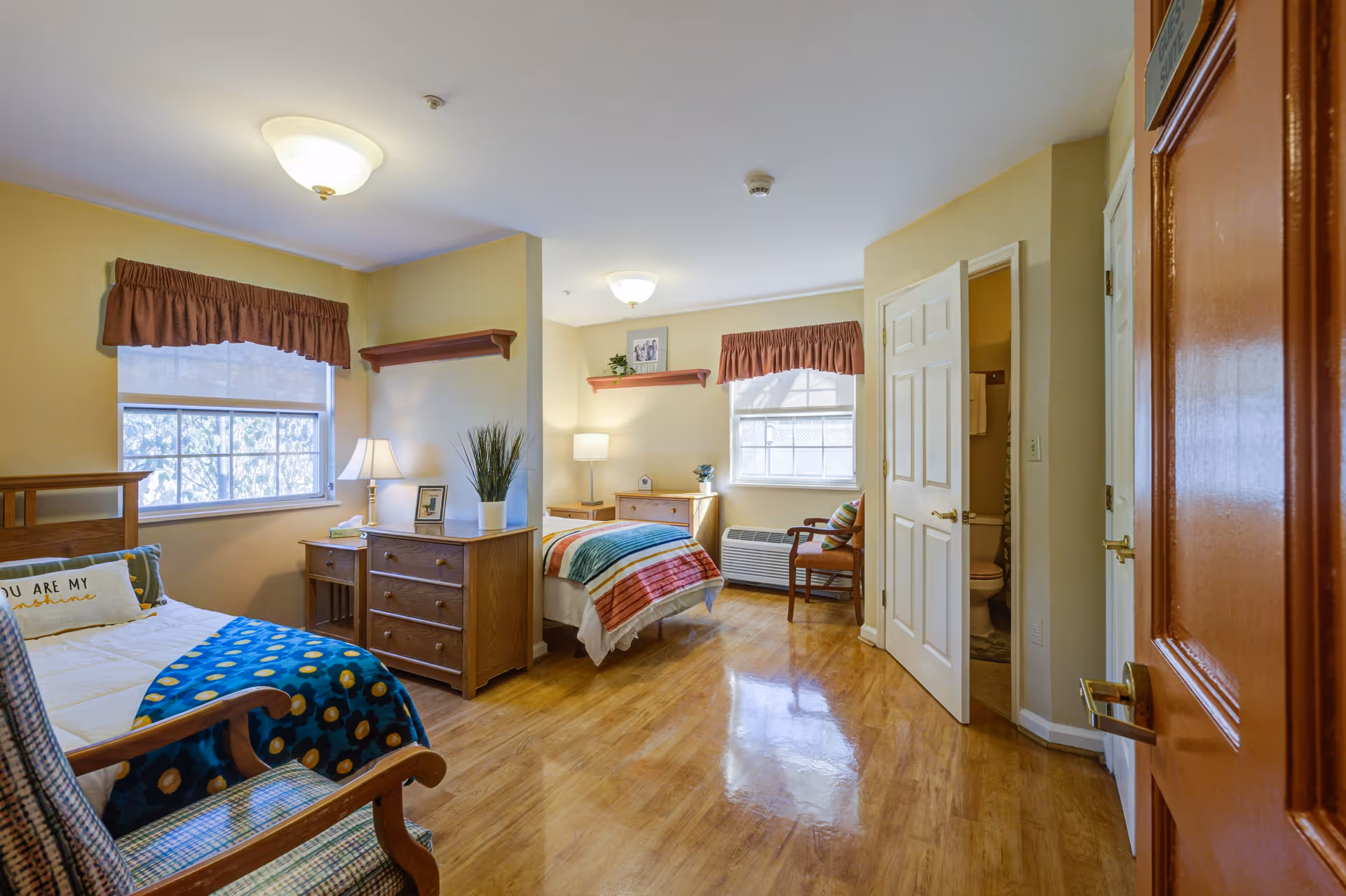 A bright and cozy bedroom in a senior living facility with two single beds, wooden furniture including dressers and nightstands, two windows with valance curtains, a wooden armchair, and an open door leading to a bathroom.