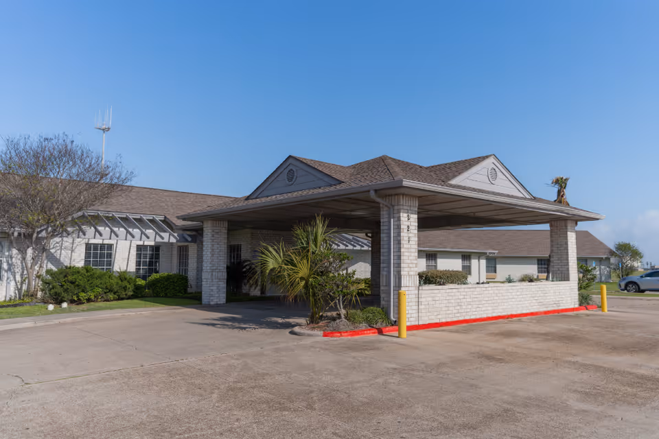 Front entrance of a single-story brick nursing facility with a covered porte-cochere and landscaping under a clear blue sky.