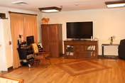 A cozy living room area with wooden flooring, a large flat-screen TV on a wooden cabinet, a small desk with a computer and chair, a wooden chair with a cushion, and a small table with decorative items. The room has beige walls and a window with beige curtains.