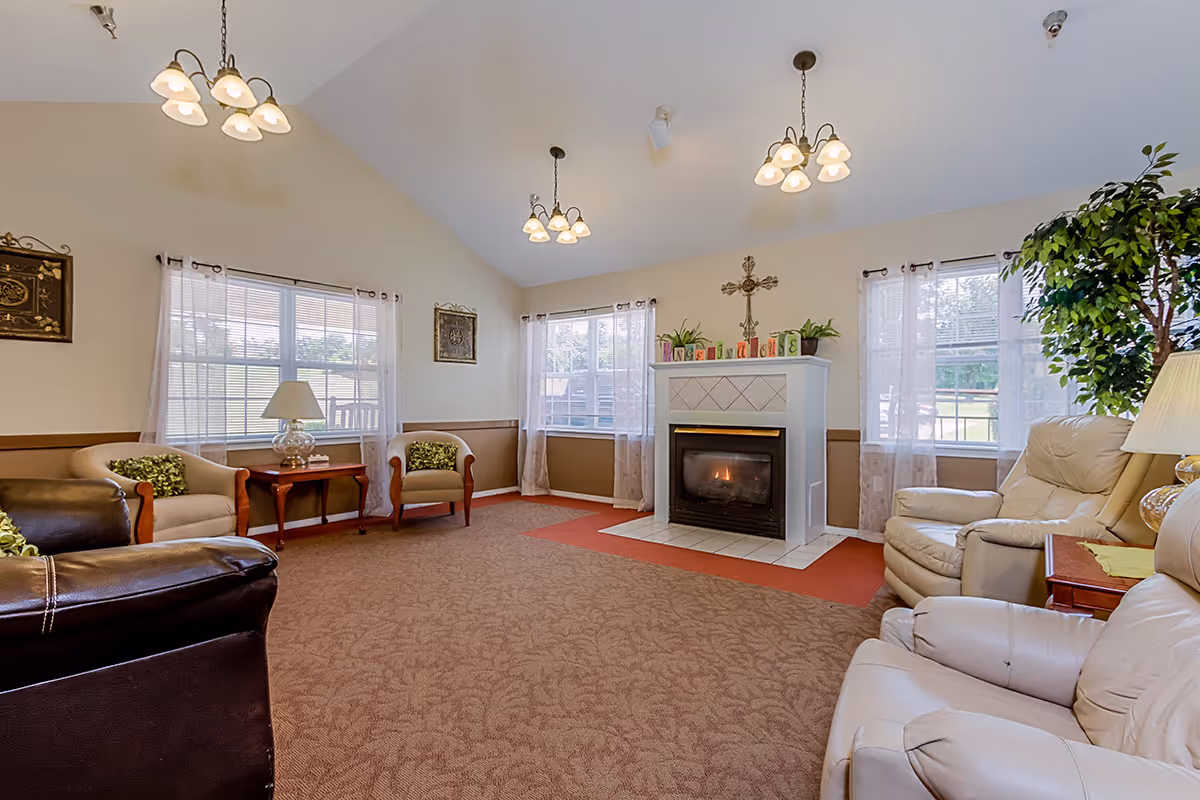 A cozy senior living facility common area with beige walls and carpeted floor. The room features a white fireplace with a lit fire, decorated with plants and colorful blocks spelling 'WELCOME'. There are multiple windows with sheer curtains allowing natural light in. Comfortable seating includes leather and fabric armchairs arranged around the room, along with side tables and lamps. The ceiling is vaulted with three hanging light fixtures.