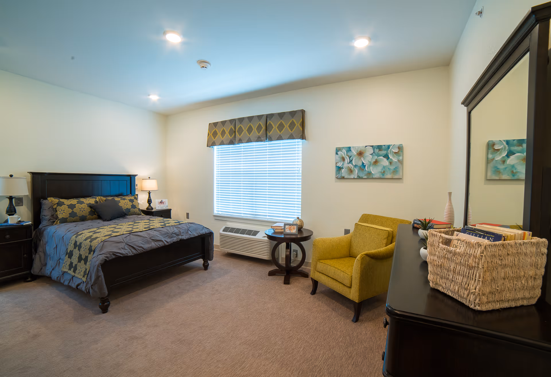 A well-lit bedroom with a large bed featuring a dark wooden frame and gray and yellow bedding. There are two bedside tables with lamps on either side of the bed. A window with blinds and a patterned valance is centered on the far wall. To the right, there is a yellow upholstered armchair next to a small round wooden table with decorative items. A dark wooden dresser with a large mirror and a basket filled with books is in the foreground on the right side. The walls are light-colored and there is a floral painting hanging above the armchair.