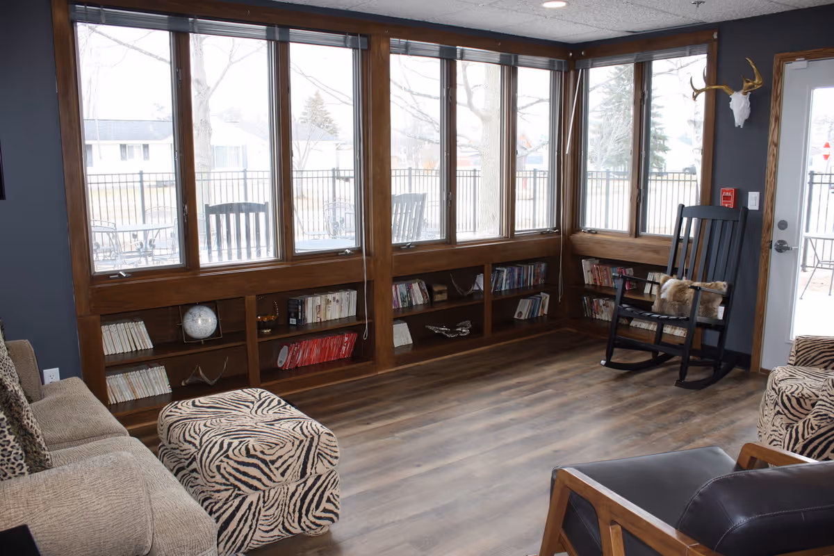 A cozy living room with large windows along two walls, wooden built-in bookshelves filled with books and decorative items, a beige sofa with patterned cushions, a zebra-print ottoman, a black rocking chair with a fur throw, and a wooden armchair with black leather cushions. Outside the windows, there is a fenced patio area with outdoor furniture.