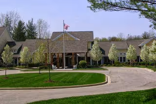Exterior view of a single-story retirement community building with a covered entrance, an American flag on a flagpole, and neatly landscaped grounds with green grass and blooming trees.