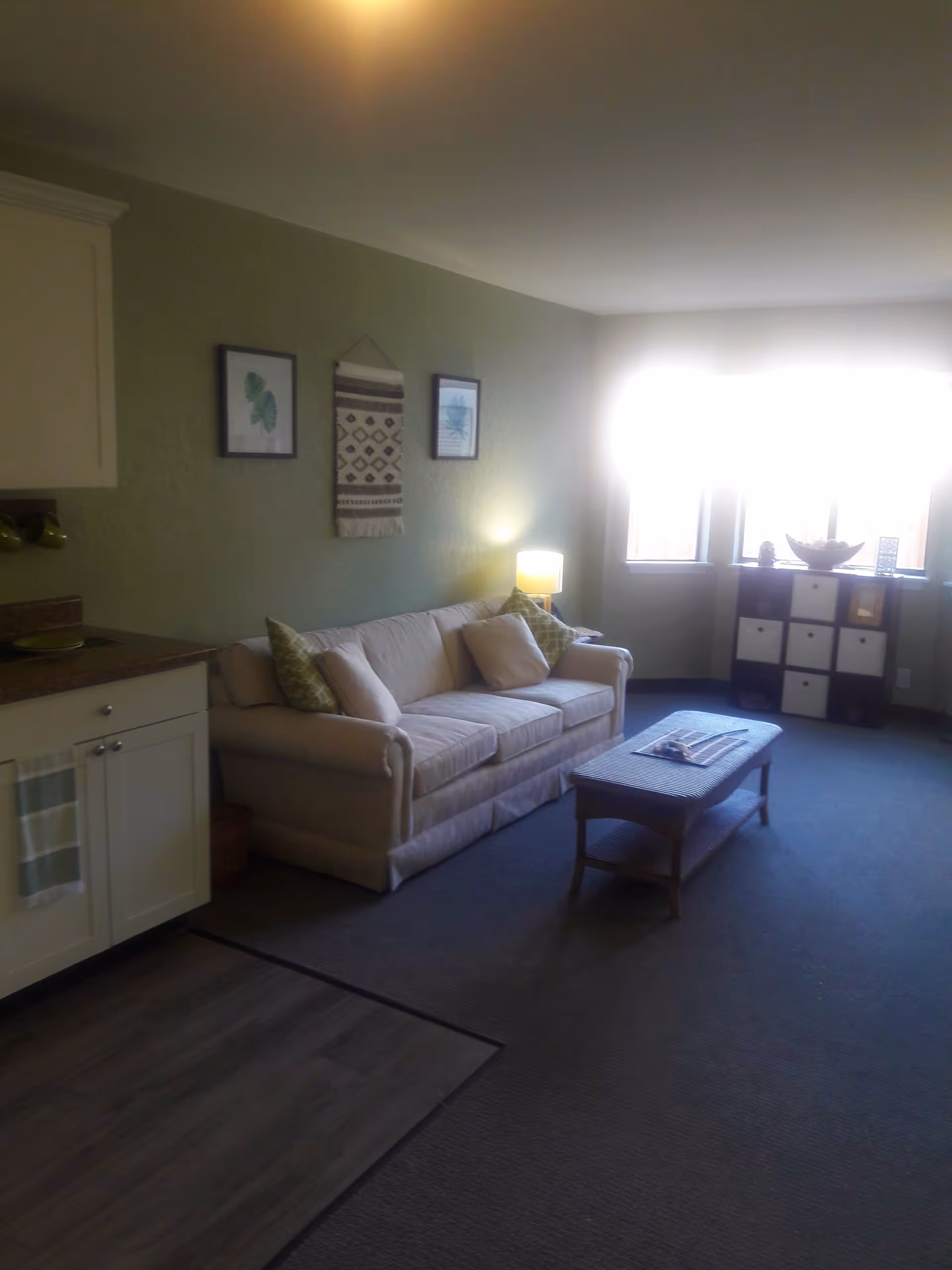 Sunlit living room with a beige sofa, coffee table, side lamp, wall art, and a small kitchenette area.