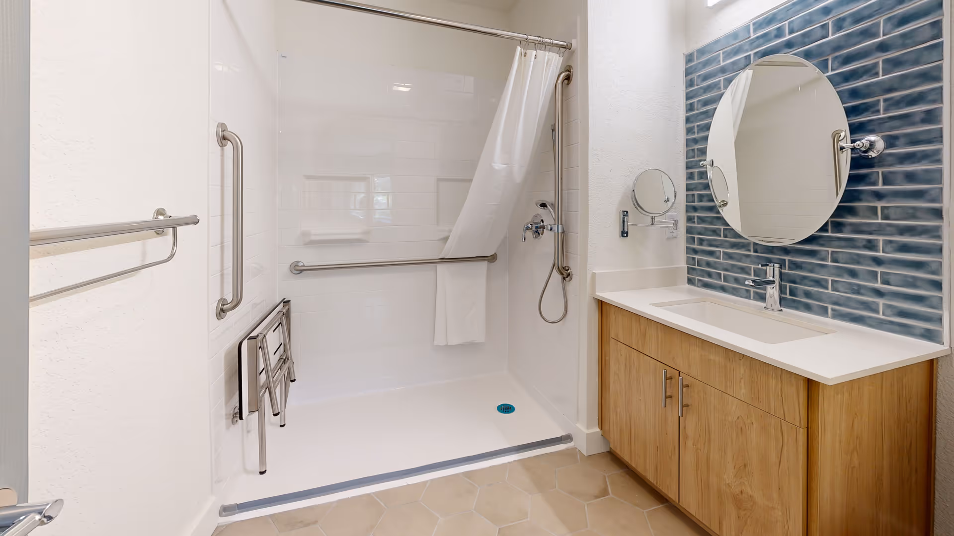 A modern bathroom featuring a walk-in shower with white tiled walls, a white shower curtain, and stainless steel grab bars. There is a foldable shower seat inside the shower. Next to the shower is a wooden vanity with a white countertop, a rectangular sink, and a round mirror mounted on a blue tiled backsplash. The floor has beige hexagonal tiles.