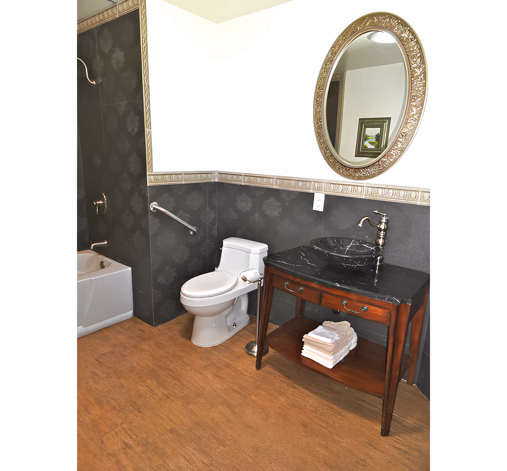 A bathroom with a white toilet, a dark wooden vanity with a black marble countertop and a round decorative mirror above it. The vanity has a vessel sink with a bronze faucet and a stack of folded towels on the lower shelf. The walls are partially tiled with dark tiles and a decorative border, and the floor has a wood-like finish. A bathtub is partially visible on the left side.