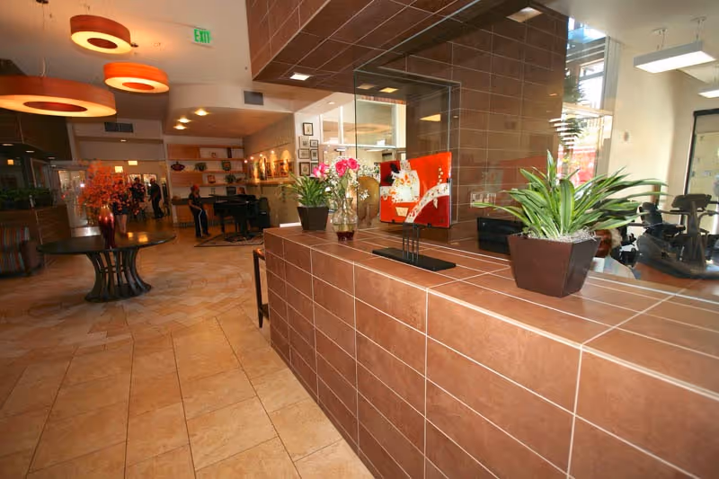 Interior view of a senior living facility lobby or common area with a tiled reception desk adorned with potted plants and decorative items. The space features warm lighting with circular ceiling fixtures, a tiled floor, and a seating area with a piano and people in the background.