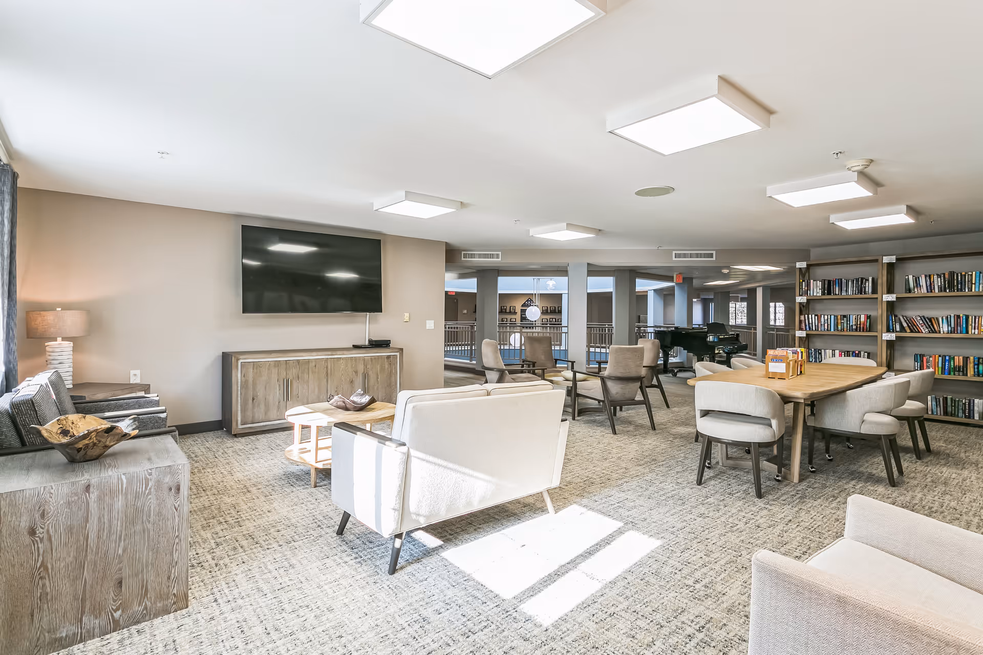 Bright communal lounge with sofas, armchairs, a large wall-mounted TV, bookshelves and a dining table.