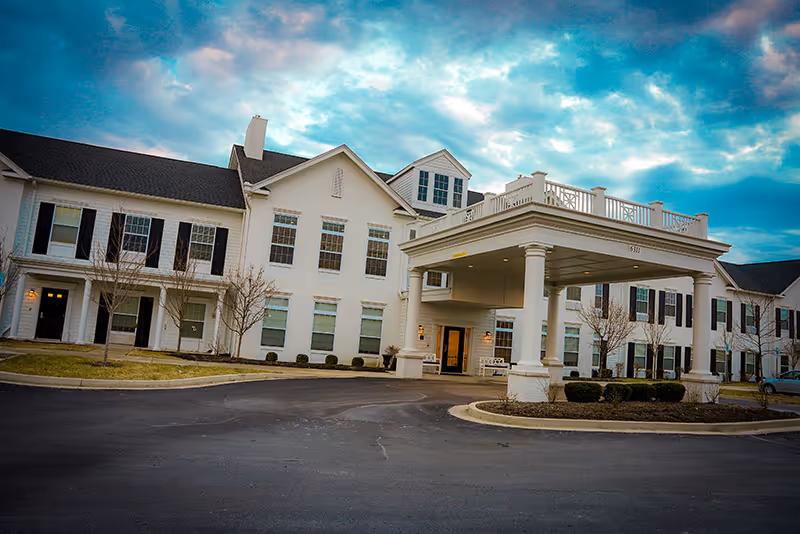 Front entrance of a large white senior living building with a porte-cochère and paved driveway under a blue sky.