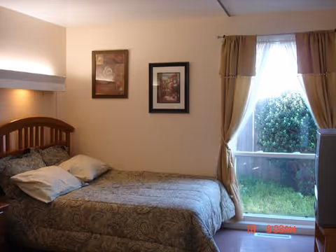 A bedroom with a wooden bed frame and a patterned bedspread with two pillows. Two framed pictures hang on the beige wall above the bed. A large window with beige curtains is on the right side, showing a green bush and grass outside. A television is partially visible on the right side near the window.
