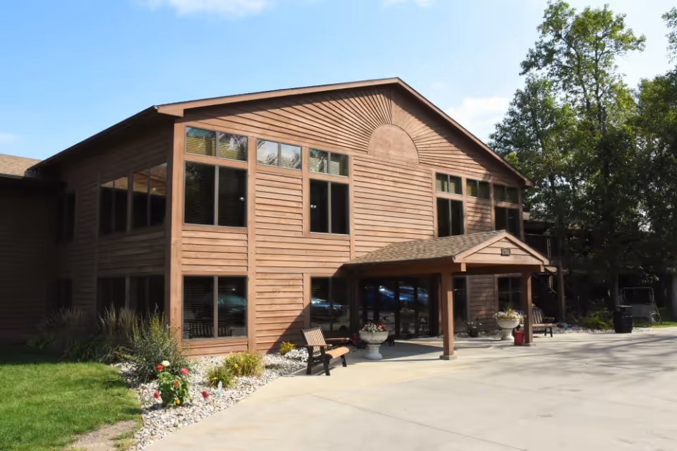 Front exterior of a two-story wood-paneled senior living building with an entrance canopy, benches, planters, and landscaping.