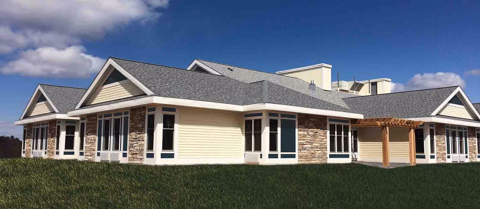 Exterior view of a single-story building with beige siding, stone accents, multiple large windows, and a gray shingled roof under a partly cloudy blue sky. The building is surrounded by green grass and features a small wooden pergola near one corner.