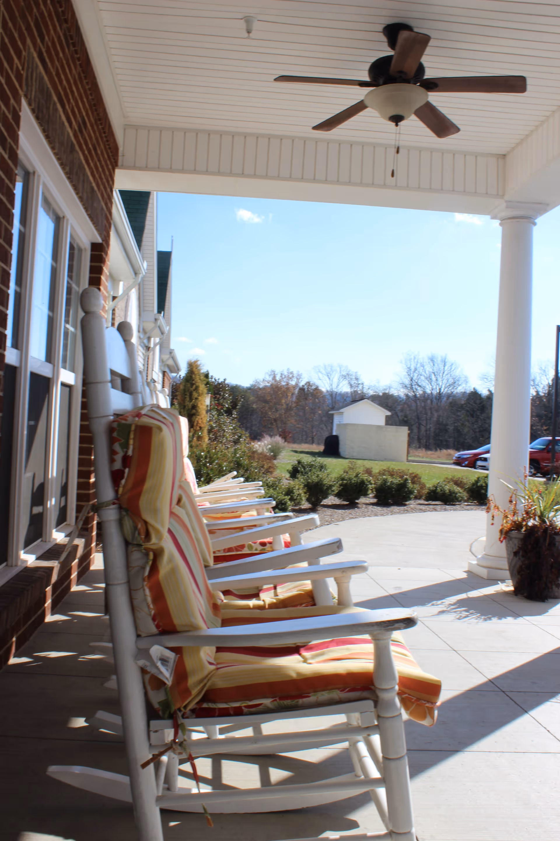 Covered front porch with a row of cushioned white rocking chairs, a ceiling fan, columns, and a view of the lawn and driveway.