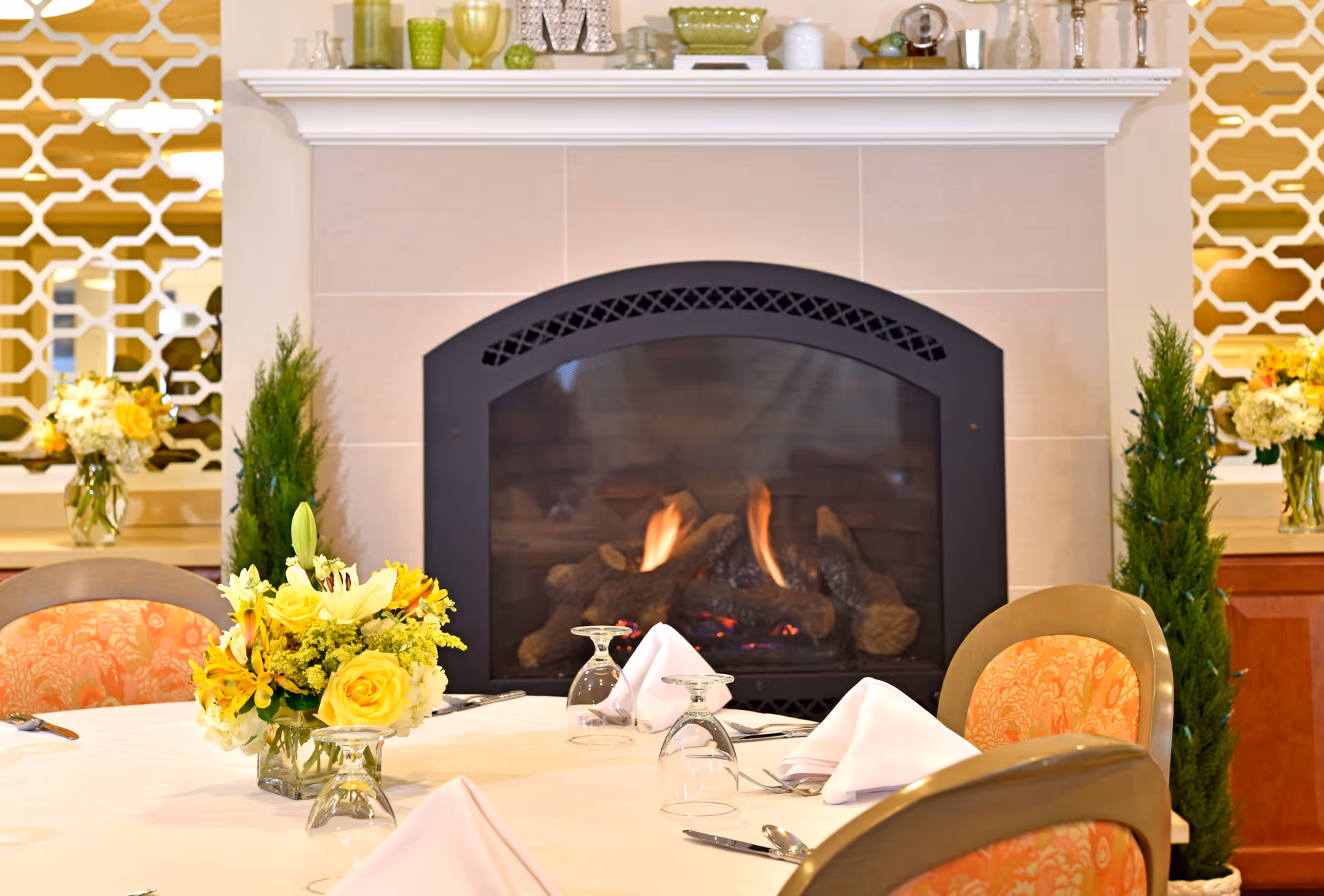 A cozy dining area with a round table set with white napkins, upside-down wine glasses, and silverware. In the background, there is a lit fireplace with a black frame surrounded by beige tiles. The mantel above the fireplace is decorated with various glassware and decorative items. On either side of the fireplace, there are green plants and floral arrangements with yellow and white flowers. The chairs around the table have orange patterned upholstery.