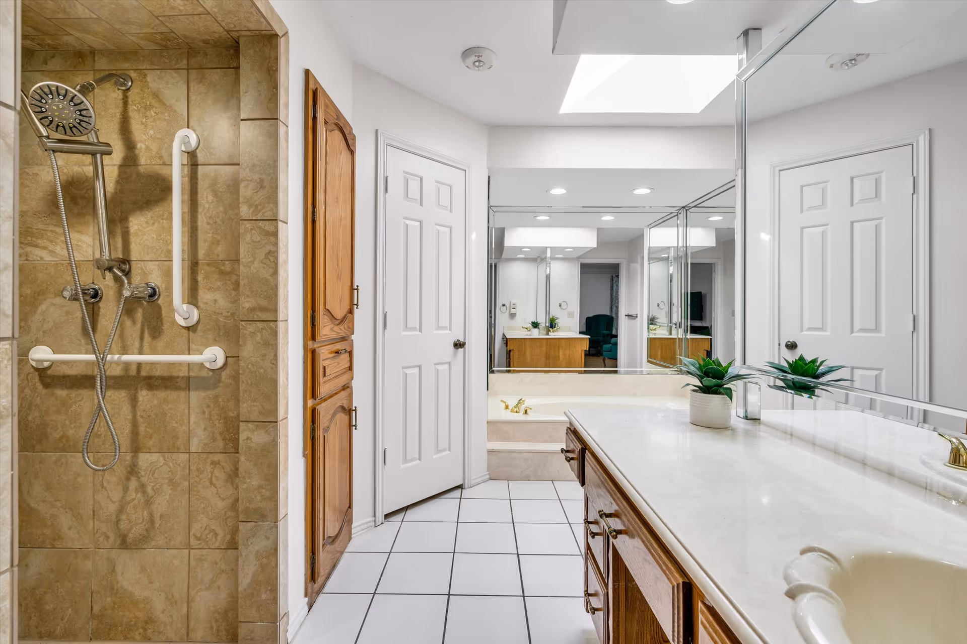Bright bathroom with a tiled walk-in shower (with grab bars), long marble countertop vanity, large mirrors, and a bathtub.